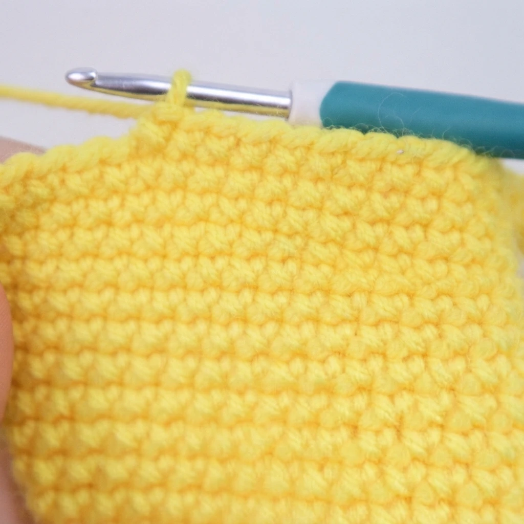 Close-up of a yellow crocheted swatch with dense single crochet stitches and a teal-handled crochet hook resting along the top edge, with a strand of yellow working yarn extending to the left.