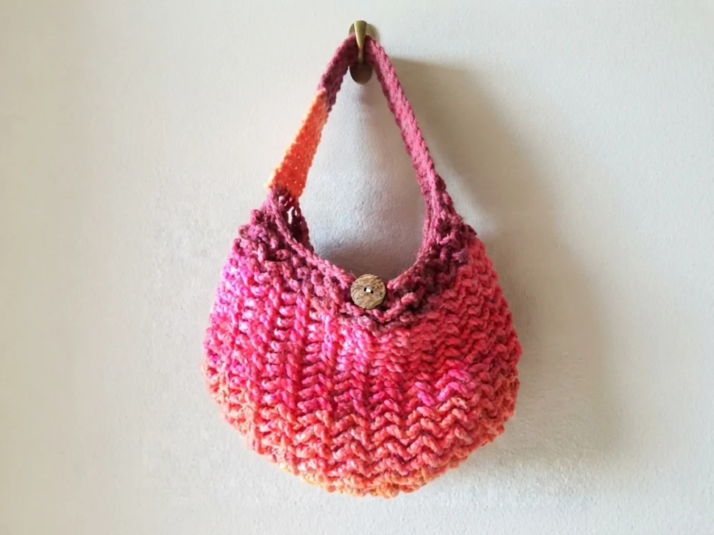 A finished crocheted market tote bag in self-striping pink, coral and orange yarn with textured V stitches, a wooden button closure and a long striped strap, hanging against a light wall.