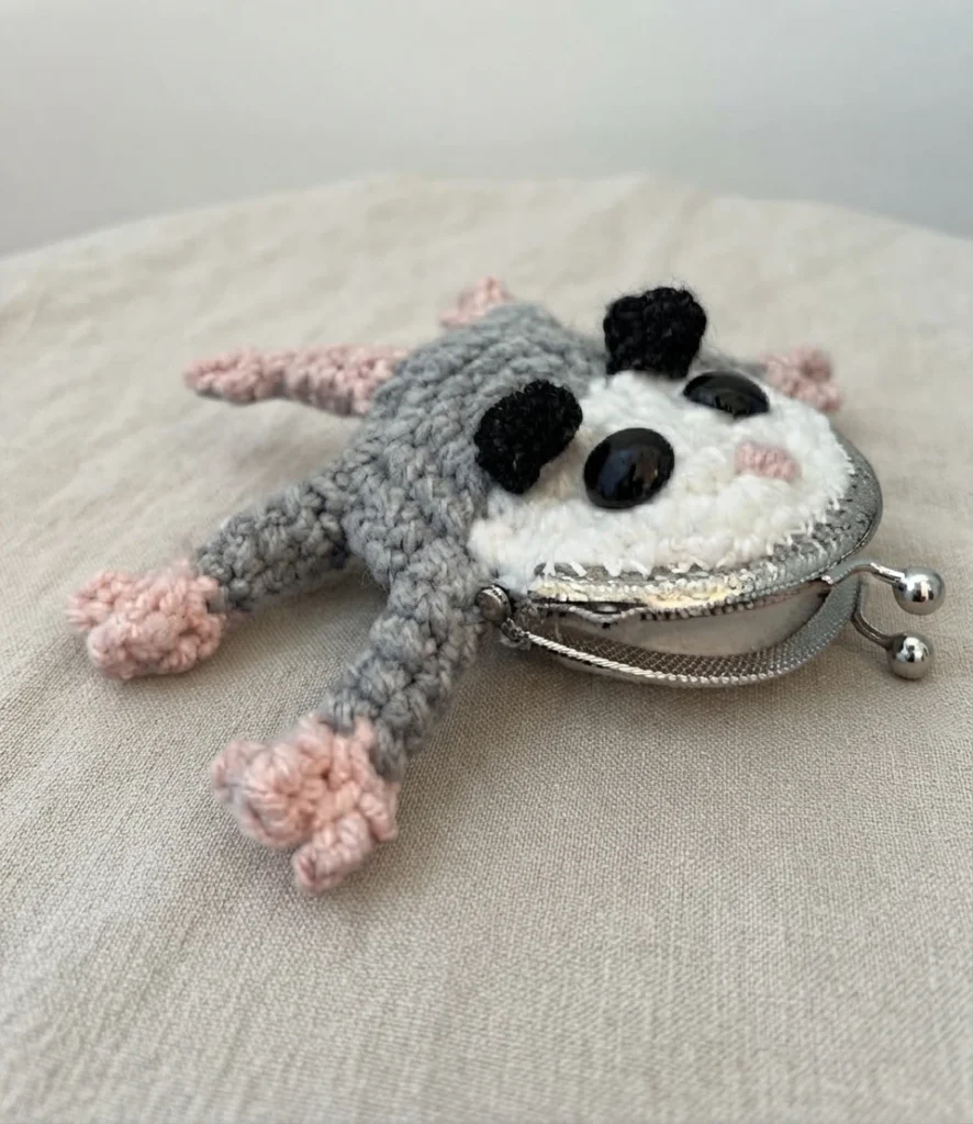 Pochette crochetée en forme d’opossum gris et blanc, avec des oreilles noires, des pattes roses et une fermeture métallique à boules.