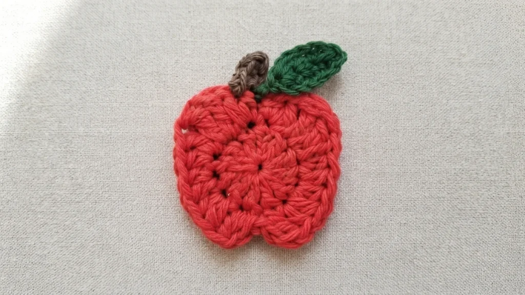 Crochet apple applique in red yarn with a small brown stem and a green leaf, lying flat on a light woven fabric background.