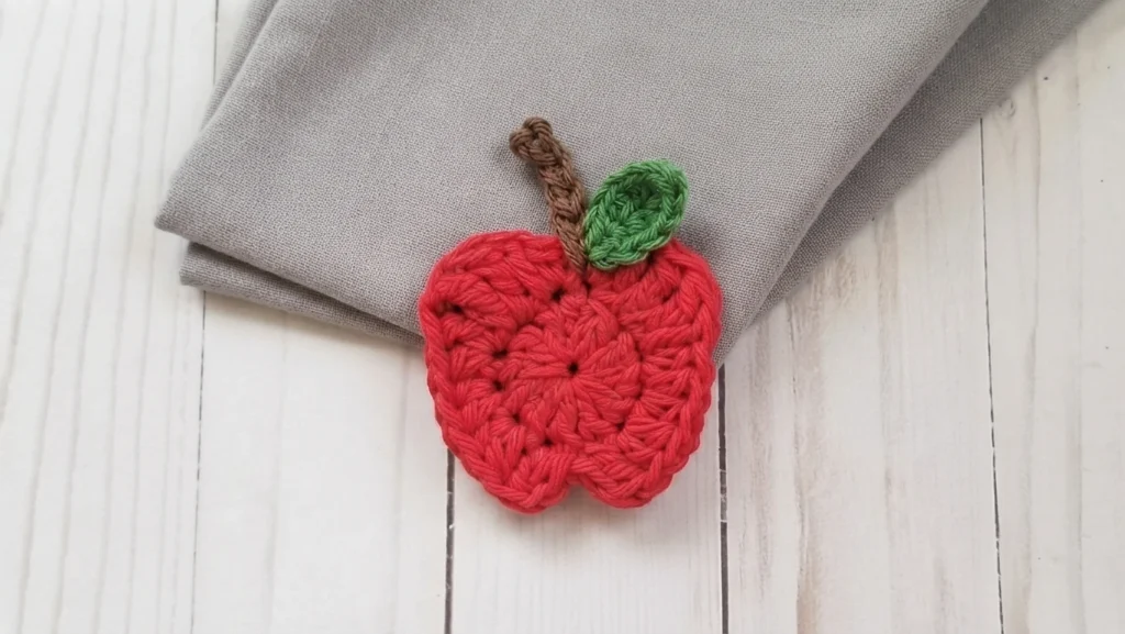 Appliquer crochet en fil rouge en forme de pomme, avec tige marron et petite feuille verte, posée sur un tissu gris plié sur une surface en bois blanc