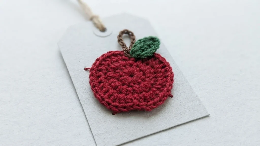 Crochet apple applique in red yarn with a brown stem and green leaf, resting on a light gray paper tag with a string loop.