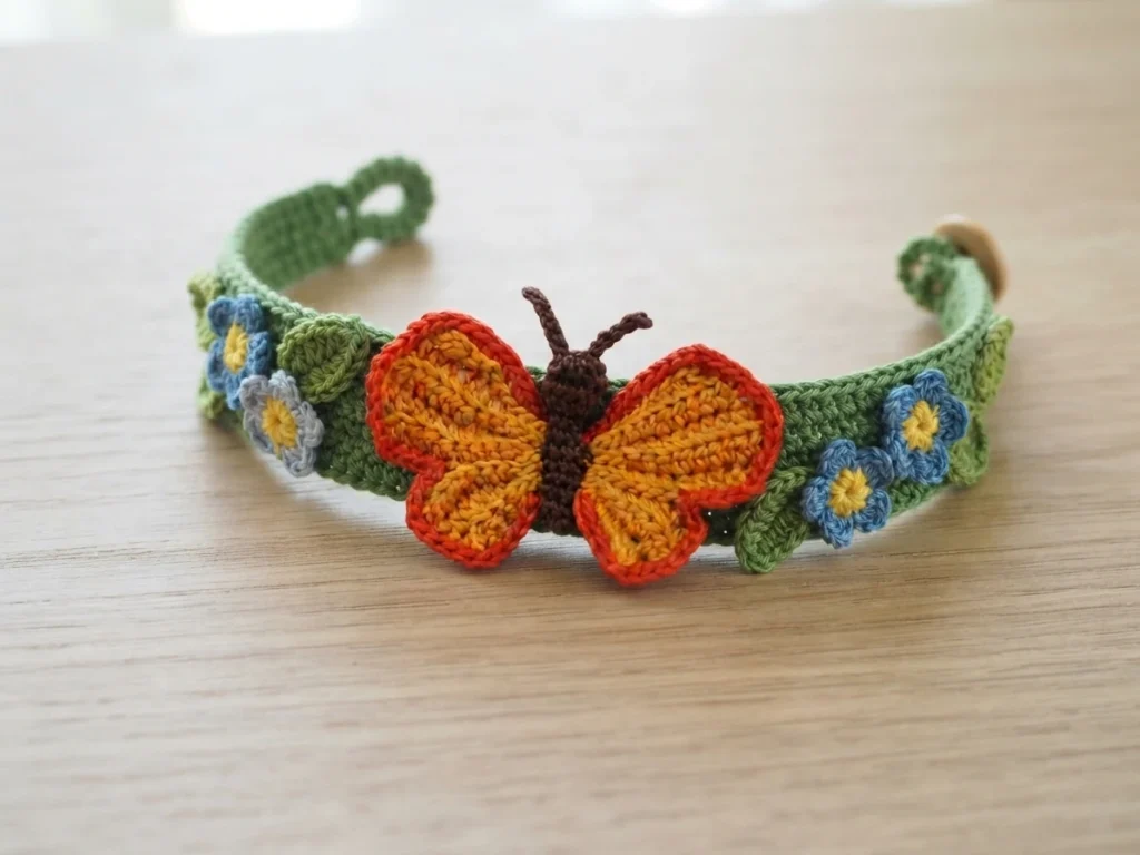 Crocheted green bracelet decorated with small blue flowers and an orange butterfly applique with brown body, closed with a button, resting on a wooden surface.