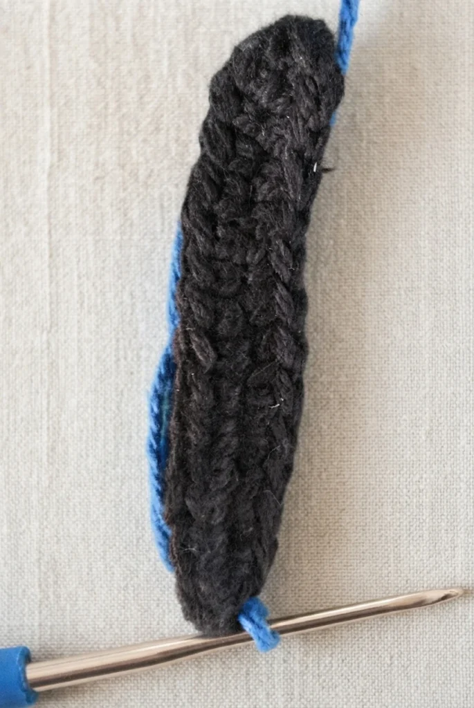 Close-up of the crochet butterfly body in dark yarn with blue yarn joined at the top and a crochet hook still holding the working loop.