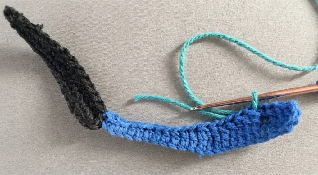 Crocheted butterfly body in dark yarn with a blue wing segment in progress, a crochet hook and teal yarn on a neutral background