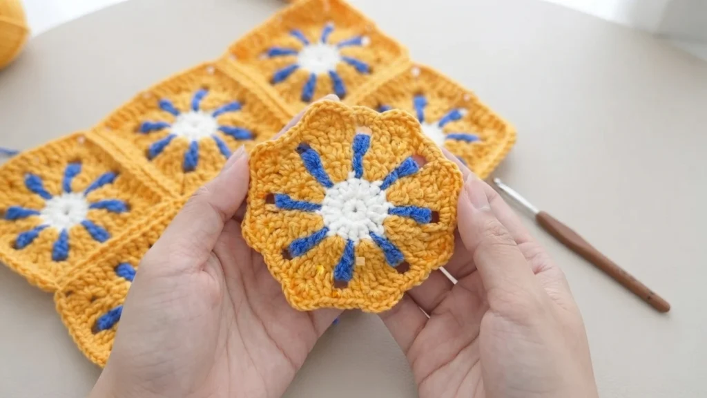 Hands holding a yellow, blue and white crocheted flower motif in progress, with joined square motifs and a crochet hook in the background.