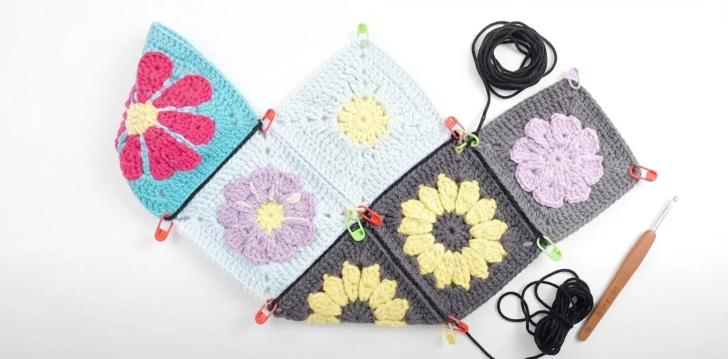 Six crochet granny squares with floral motifs pinned in a zigzag layout, joined at the edges with black yarn, with a crochet hook and yarn balls beside them.