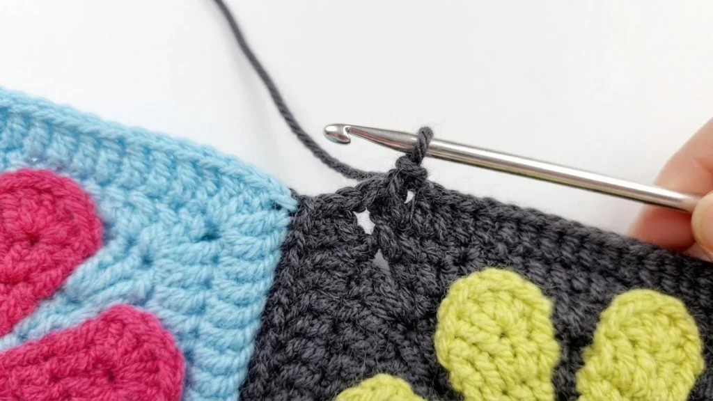 Close-up of a metal crochet hook pulling dark yarn through the edges of two crocheted granny squares, with a hand holding the hook; visible pink and lime-green flower motifs on blue and dark grey squares.