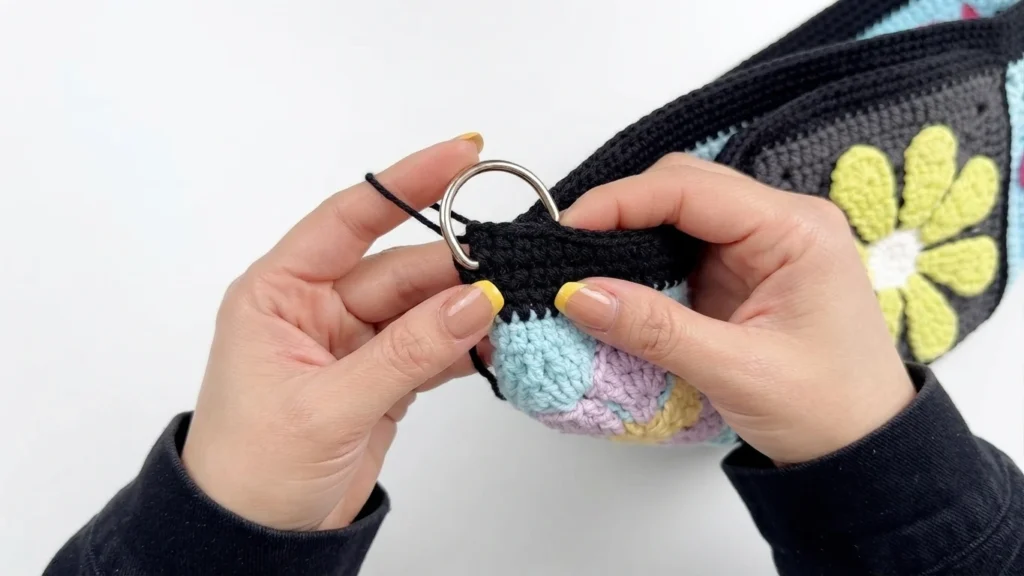 Hands attaching a silver metal ring to the black crocheted top edge of a colorful granny square bag
