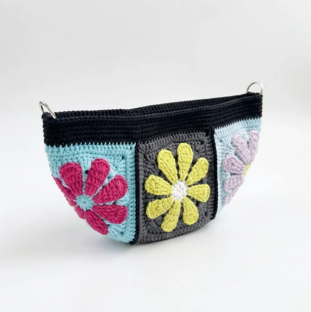 Crocheted granny square bag opening with black top edge, pink, lime green and lavender flower motifs, and two small metal D-rings on a white background.