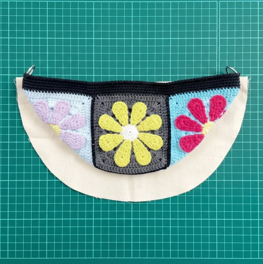 Flat crochet bag panel with three flower granny squares resting on a cut cream lining fabric on a green cutting grid mat