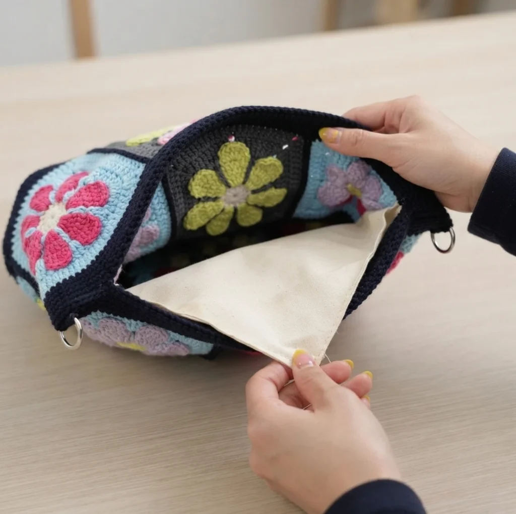 Hands tucking a cream fabric lining inside an open crochet granny square crossbody bag with flower motifs