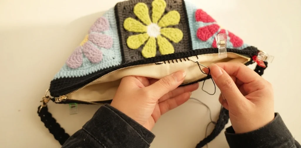 Hands hand sewing a cream lining into the open top of a floral granny square crossbody bag held with sewing clips.
