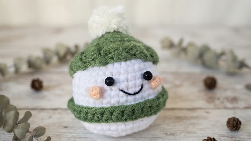 White crocheted ornament with a smiling face, green bands and a fluffy white pompom, displayed on a wooden surface.