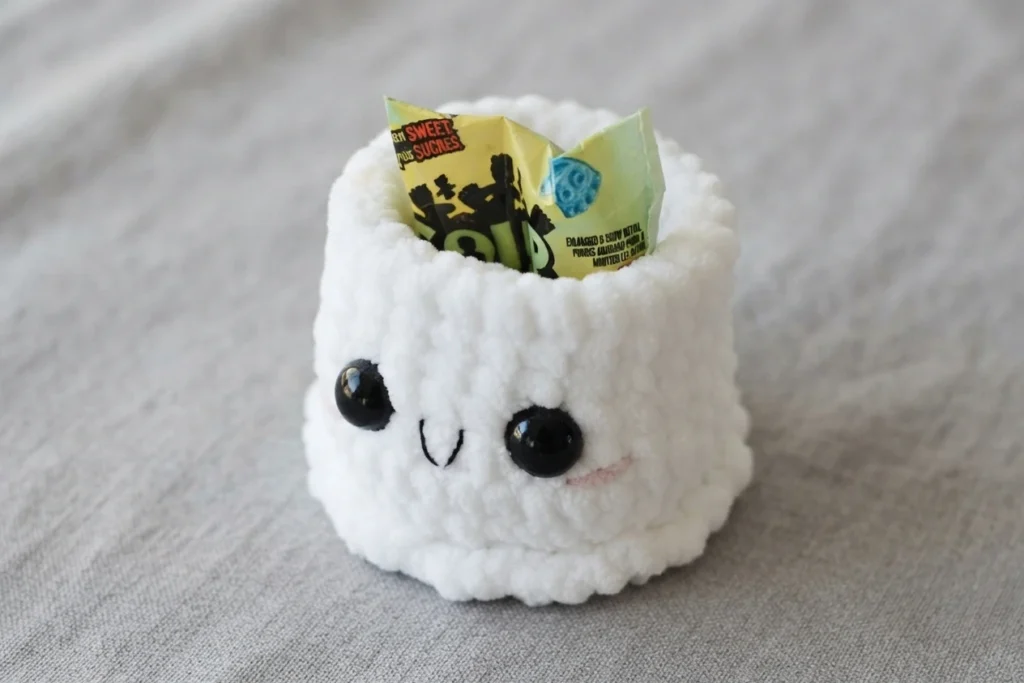 Small white crocheted ghost gift buddy with black plastic safety eyes, an embroidered smile and pink blush cheeks, holding a yellow candy wrapper sticking out of its open top, photographed on a gray fabric surface.