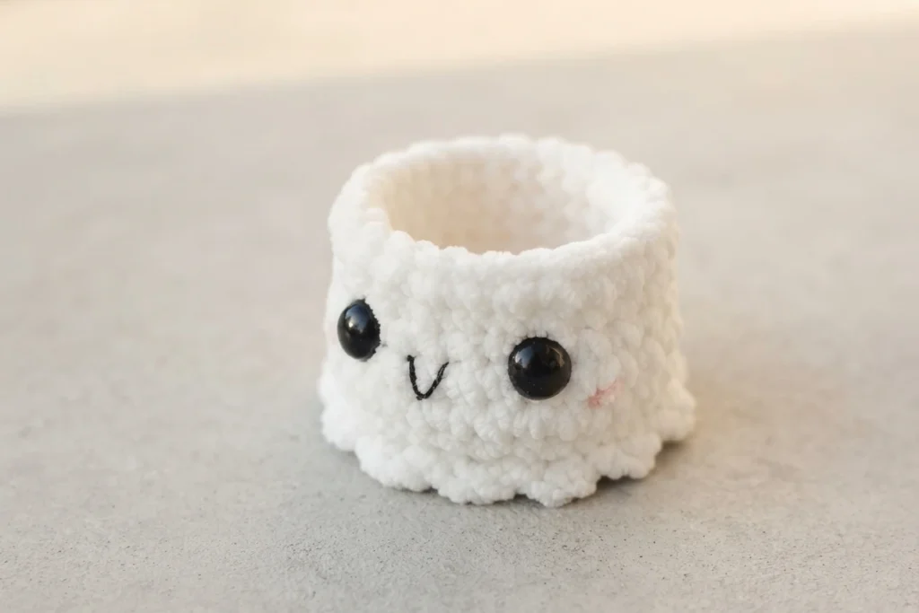 White crocheted ghost cup with black safety eyes, a stitched smile and a small pink cheek, sitting empty on a grey speckled surface