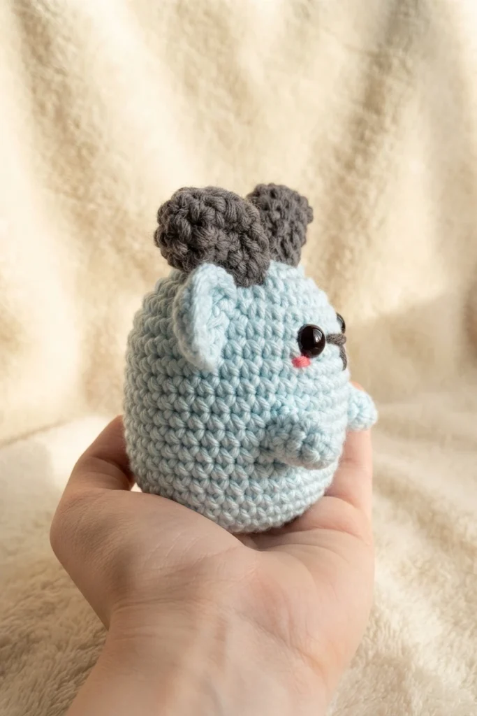 Light blue crocheted amigurumi goat with gray horns, black safety eyes and pink cheeks, held in a hand against a cream fabric background