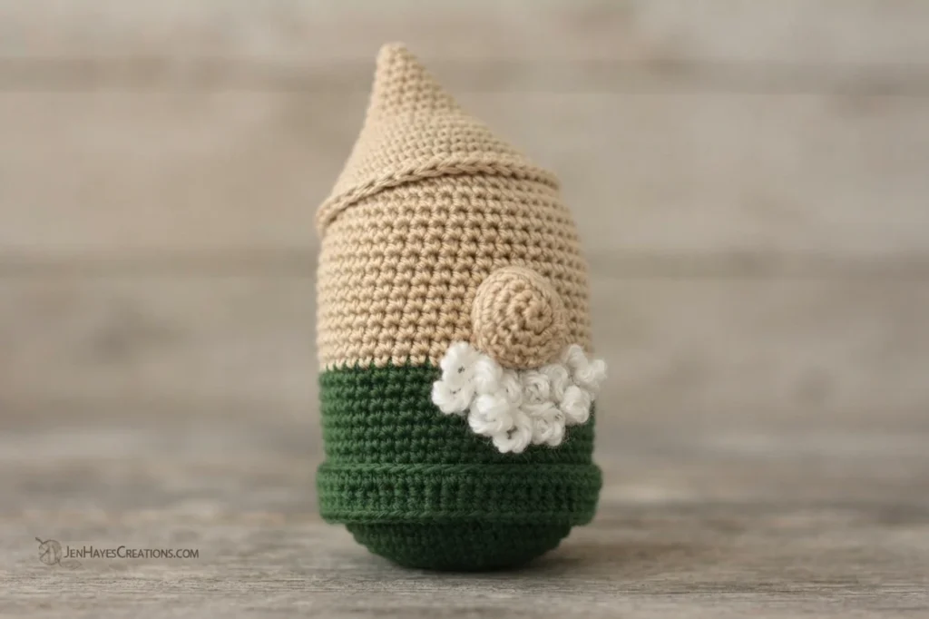 Crocheted gnome body in tan with a green tunic, a tan spiral nose and a small white fuzzy beard, standing on a light wooden surface with a small watermark in the lower left corner.