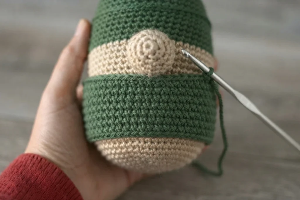 Main tenant le corps d'un gnome crocheté à l'envers, avec une partie supérieure verte, une partie inférieure beige, un petit nez en spirale beige et un crochet métallique piquant dans une rangée verte en cours de travail.