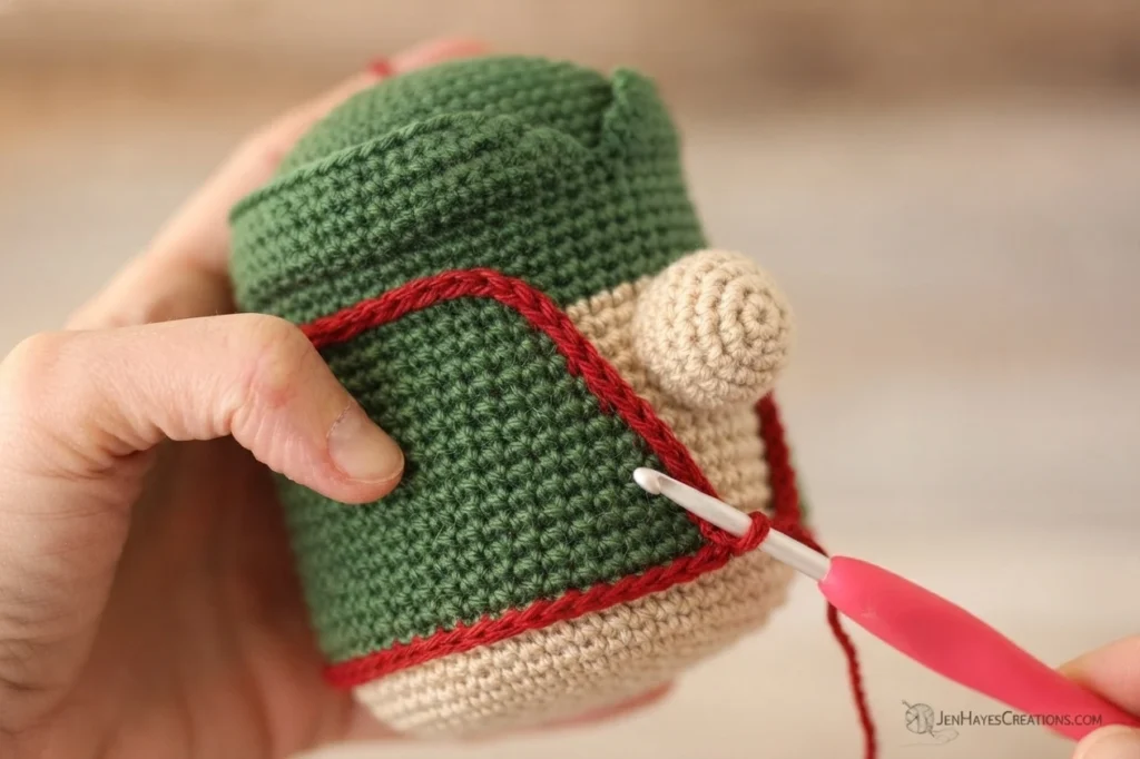 A hand holds the upside-down crocheted body of a Christmas elf gnome, showing a green tunic with a red slip-stitch trim worked along its edge and a beige spiral nose, with a pink-handled crochet hook working the red yarn.