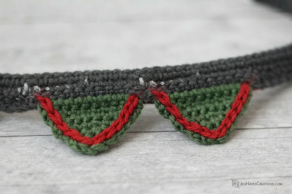 Close-up of a gray crocheted band with two green pointed collar sections edged in red surface crochet, resting on a light wood surface, with a small JenHayesCreations watermark in the lower right corner.