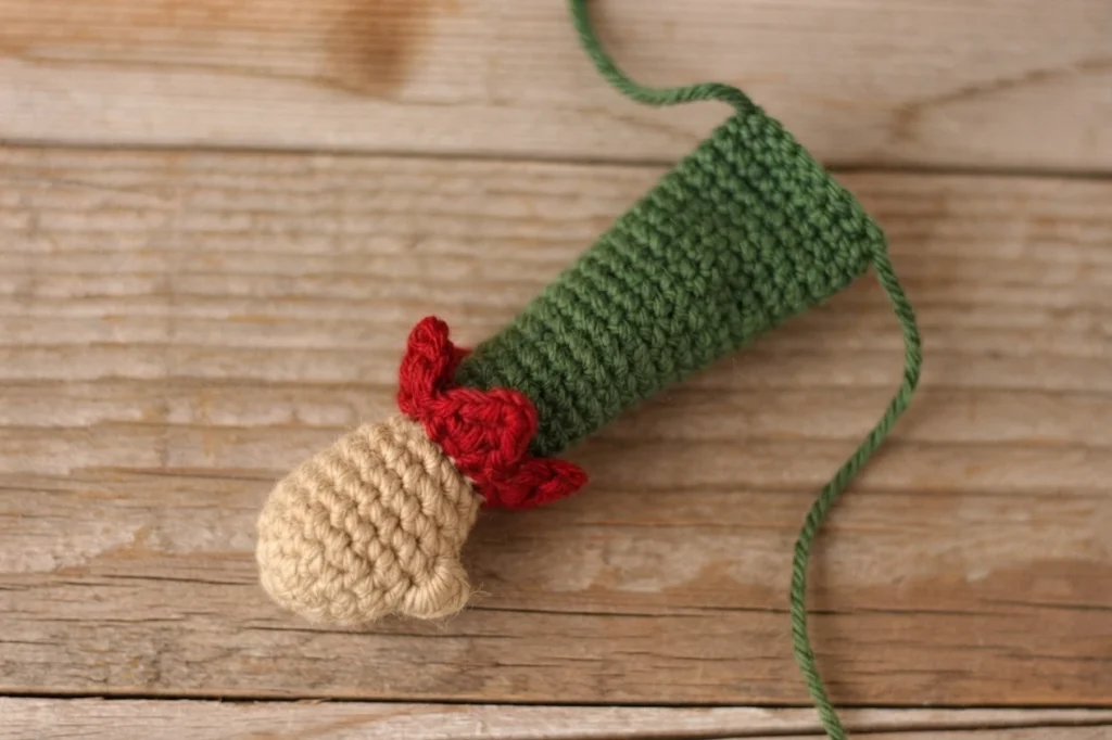 One crocheted elf arm lying on a wooden surface, with a stuffed tan hand, a small red ruffled cuff and a green tube arm, plus a long green yarn tail.