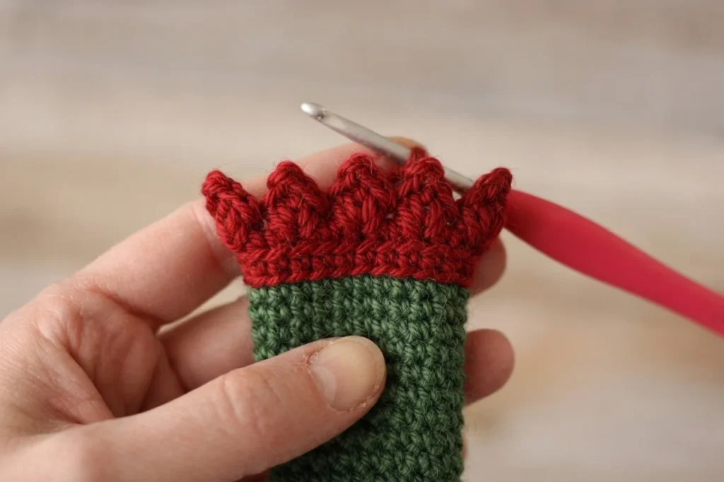 Hand holding a small green crocheted tube with a red pointed cluster edging worked along its upper edge, a metal crochet hook with a red handle resting in the stitches.