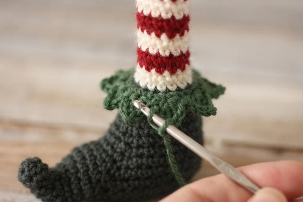 Close-up of a crocheted elf shoe in charcoal grey with a dark green ruffled cuff, a red and white striped leg above it, and a hand holding a metal crochet hook inserted into the cuff stitches.