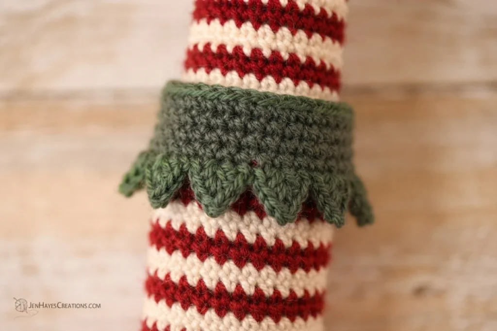 Close-up of a red and white striped crocheted leg with a dark green scalloped cuff worked around the top, photographed against a light wood surface.