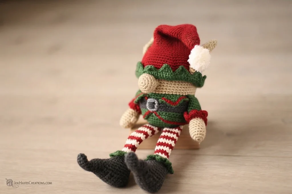 Crocheted Christmas elf gnome sitting on a light wood floor, wearing a red hat with a white pom pom, a green zigzag collar, a green body with red trim, a grey belt with a buckle, red and white striped legs and dark grey curled shoes.