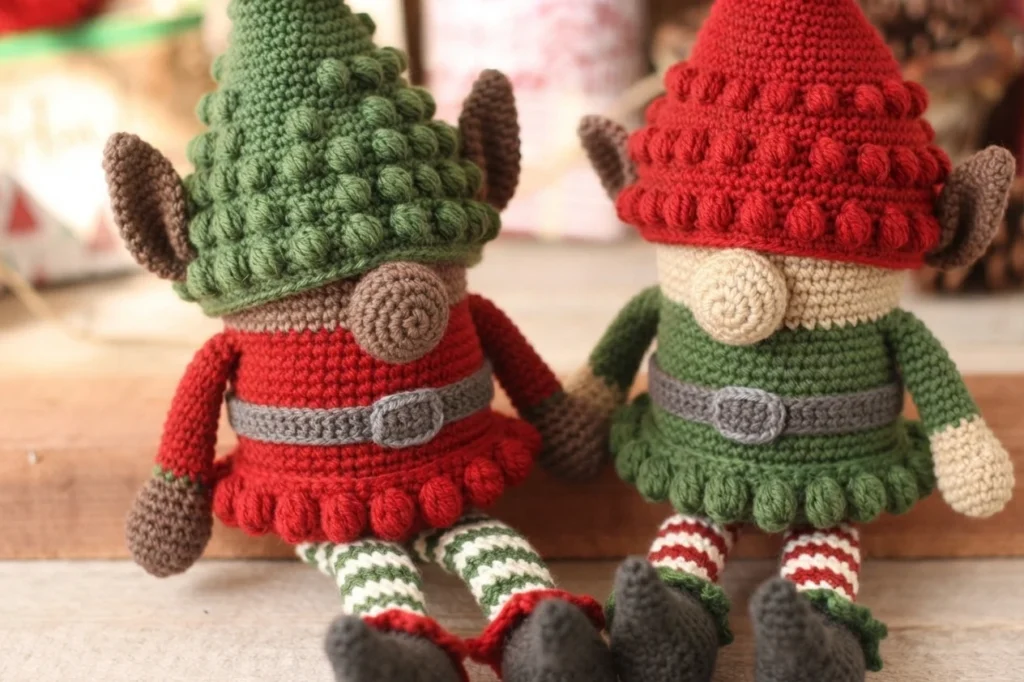 Two crochet elf gnomes sitting side by side on a wooden ledge, wearing bobble-textured pointed hats and belted tunics with striped legs and dark boots, against a soft blurred background.