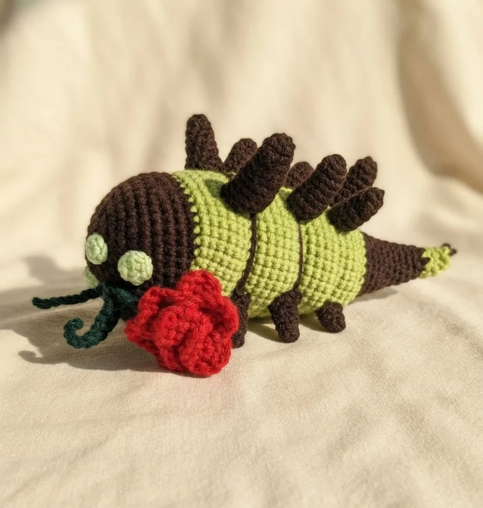 Crocheted worm amigurumi with a brown head, two pale green round eyes, a segmented light green body with dark brown back spikes and small legs, a dark green tendril, and a red crocheted rose lying beside it on cream fabric.