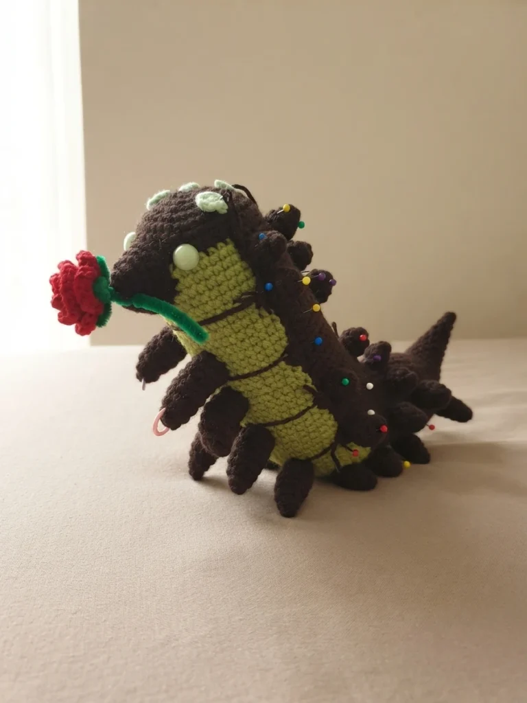 Crocheted worm amigurumi in progress with a dark brown back covered in sewing pins, a light green striped body, pale green eyes, small brown legs and a red crocheted rose near its mouth, resting on a pale surface.