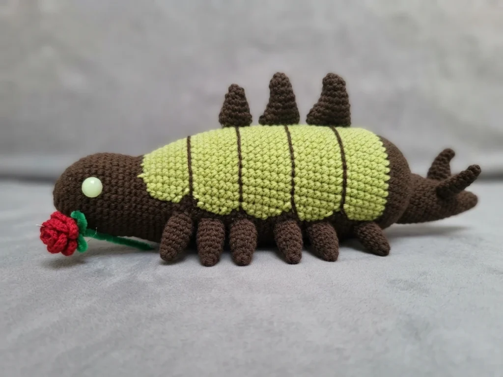 Crocheted worm toy with a dark brown head, pale green striped mid-body, brown spikes along the back and small brown legs, holding a red crocheted rose at its mouth, photographed on a light gray surface.