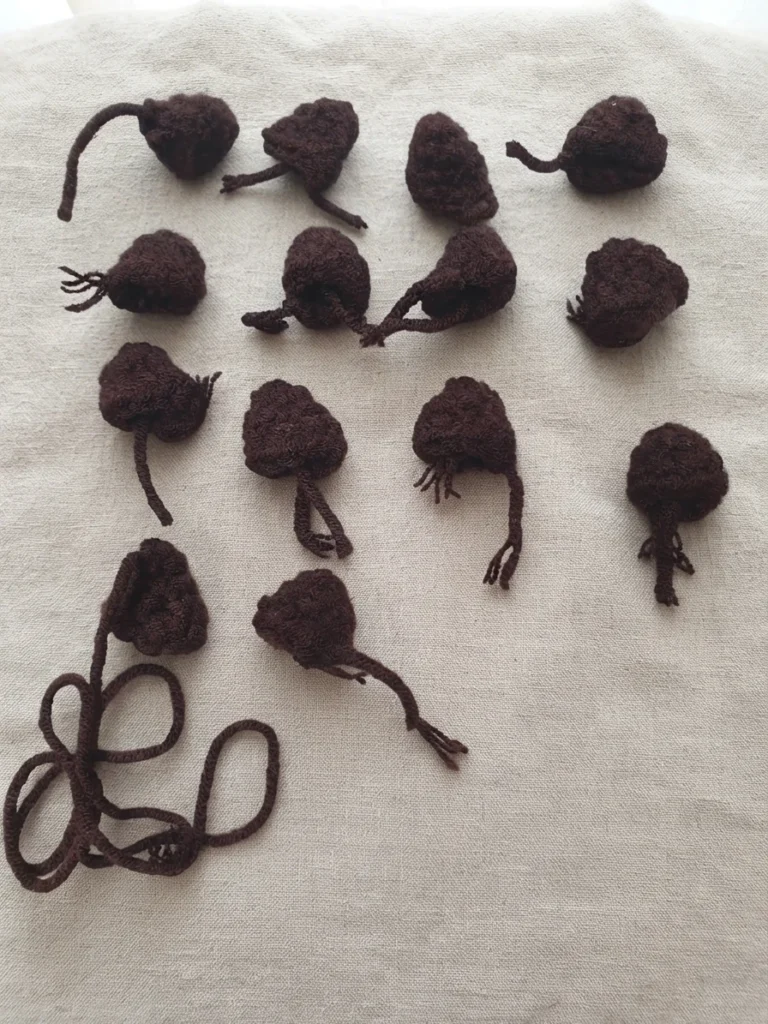 Fourteen small dark brown crocheted leg pieces with yarn tails laid out on a light beige fabric background, with a loose strand of brown yarn coiled at the lower left.