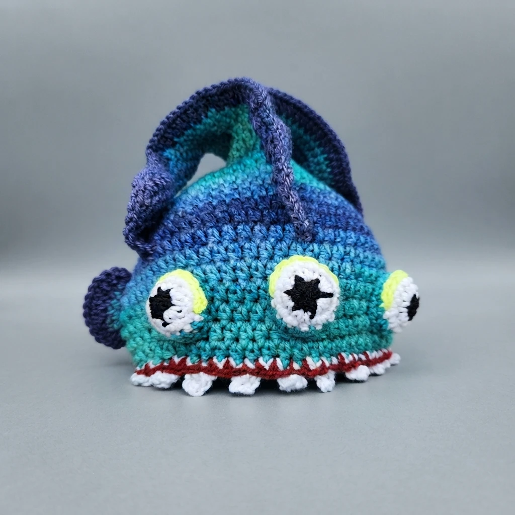 A finished crocheted abyssal eel hat in blue and teal yarn with a tall purple dorsal fin, white eyes with black star pupils and yellow eyelids, and a red gum line with white teeth