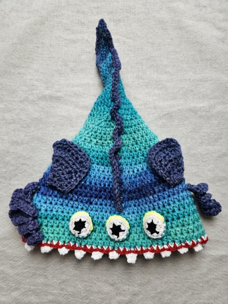 Crochet eel hat with a teal and blue striped body, purple fins, three yellow eyes with black star pupils, and a red and white toothy mouth trim