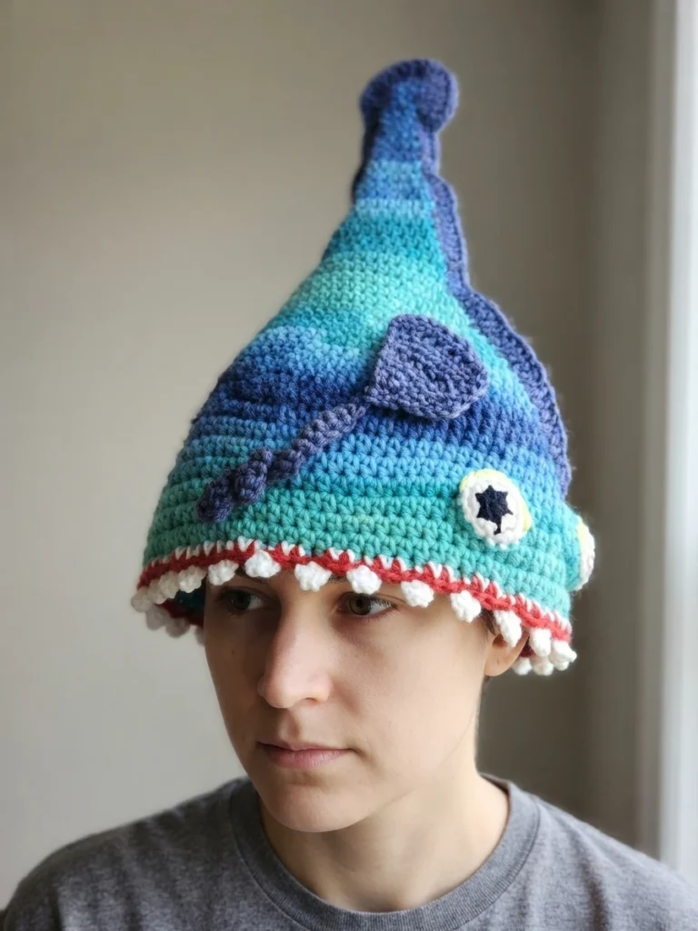 Person wearing a finished crocheted eel hat in blue and turquoise with a purple fin, an appliqued eye with a black star pupil, and a red brim edged with white teeth.