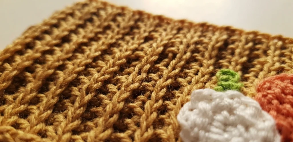 Close-up of the tan Tunisian knit ribbing of a crocheted cup cozy, with a white crocheted pumpkin motif and part of an orange pumpkin motif at the lower right edge.