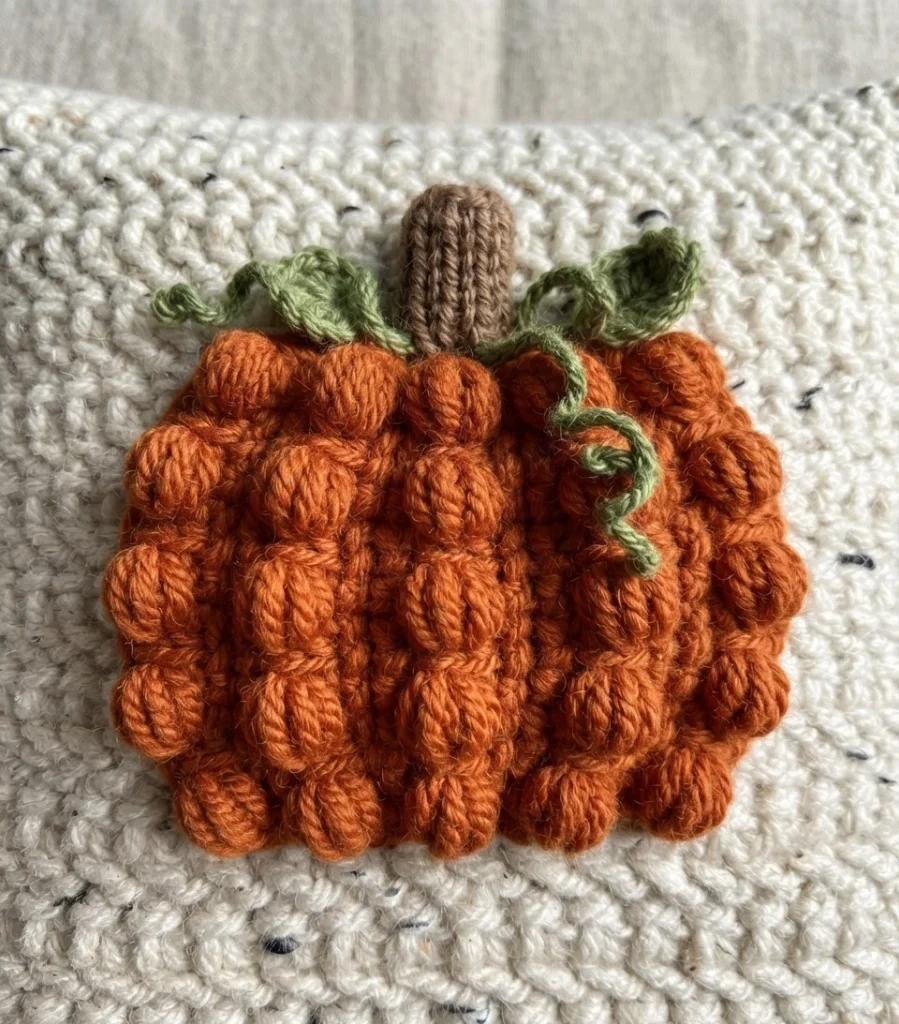 Close-up of a raised orange crochet pumpkin with a brown stem and green vines on a flecked ivory crocheted cushion.