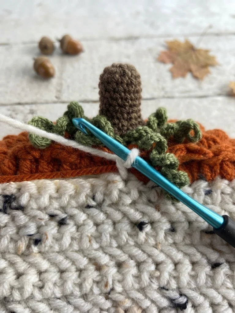Close-up of a blue crochet hook working white yarn beside orange yarn on an ivory crochet panel, with a brown pumpkin stem and green vines visible.