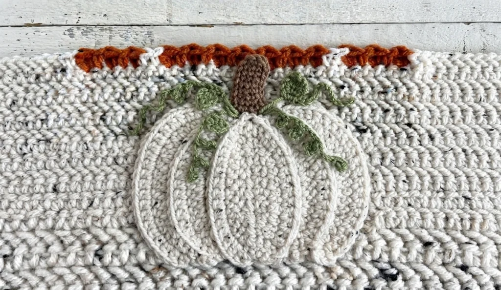 A cream tweed crochet pillow with an outlined cream pumpkin, green vines, a brown stem, and an orange border.