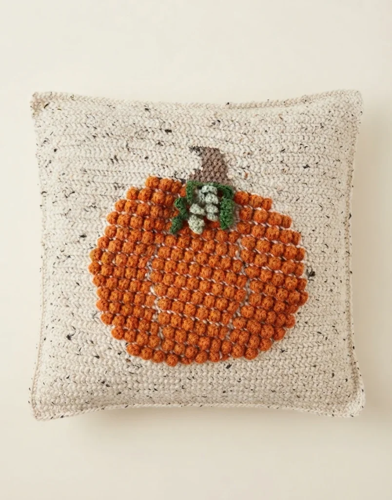 Coussin carré au crochet, en fil ivoire chiné, orné d’une citrouille orange en relief avec une tige brune et des détails verts.