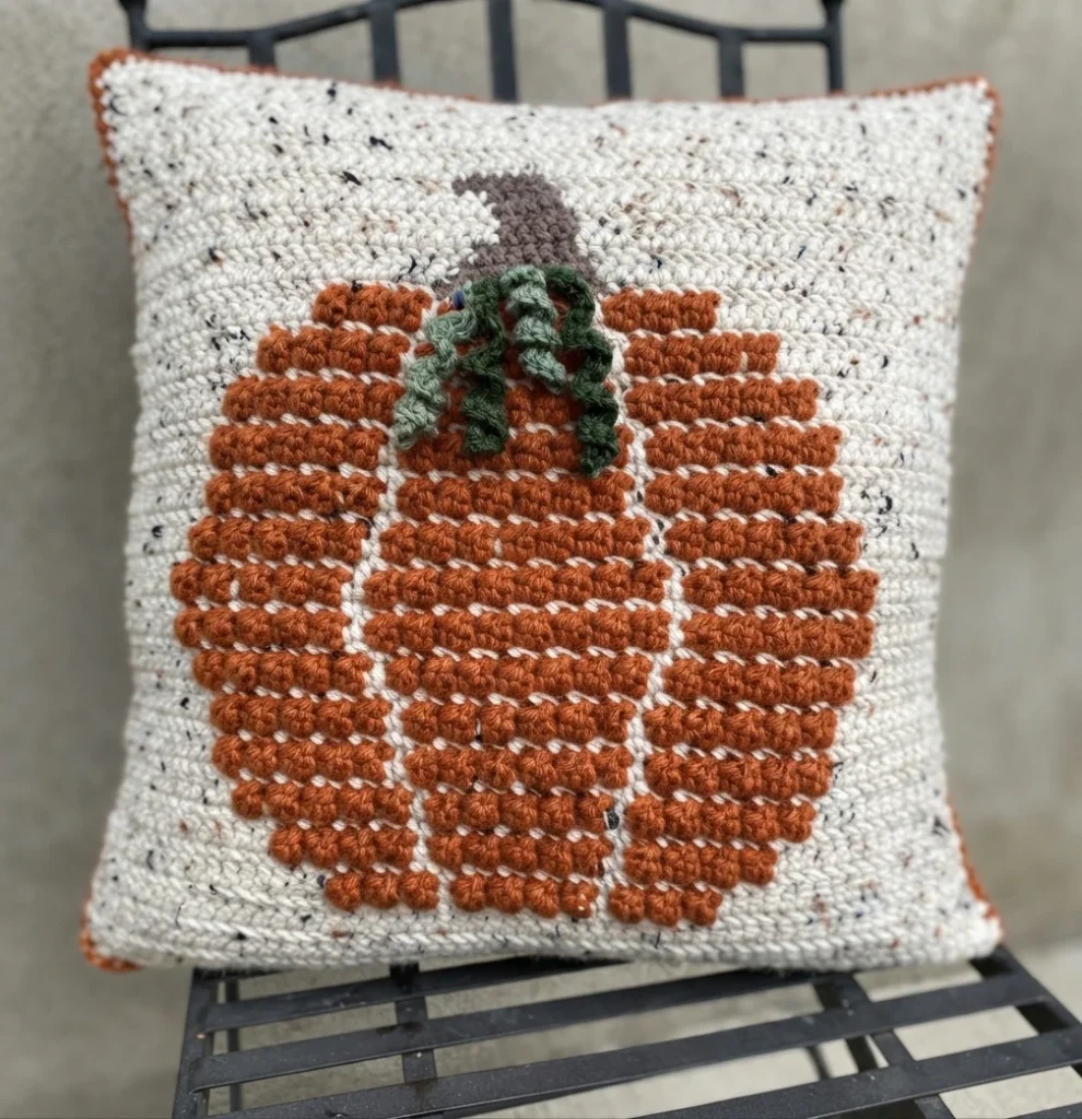 Square crocheted pillow with a raised orange pumpkin, green vines, a brown stem, and an ivory flecked background with an orange border, resting on a metal chair.