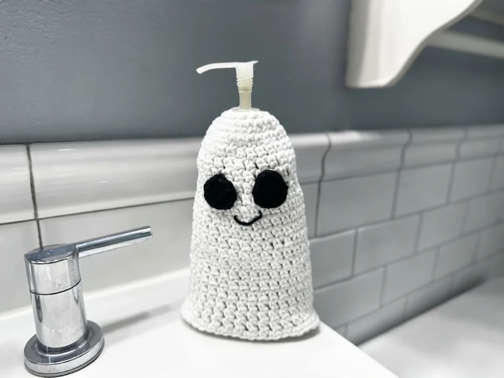White crocheted ghost soap cover with two black oval eyes and a small embroidered smile, slipped over a pump soap bottle on a white counter, beside a chrome faucet handle against white subway tiles.