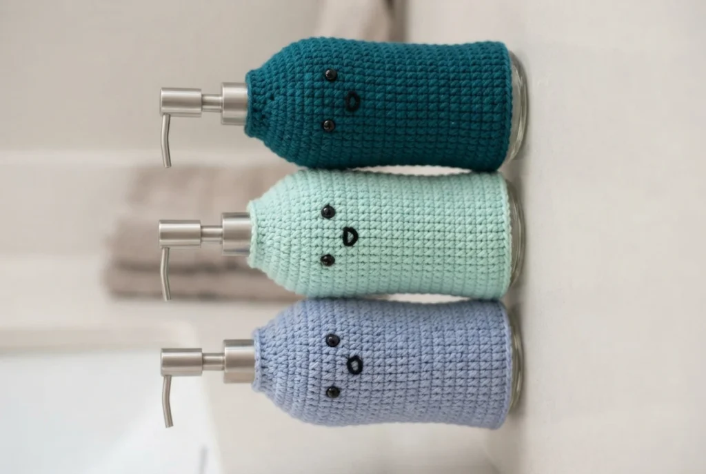 Three glass soap dispenser bottles in a row, each fitted with a crocheted sleeve in teal, mint green and light lavender, with black bead eyes and a small embroidered mouth, photographed against a pale surface.