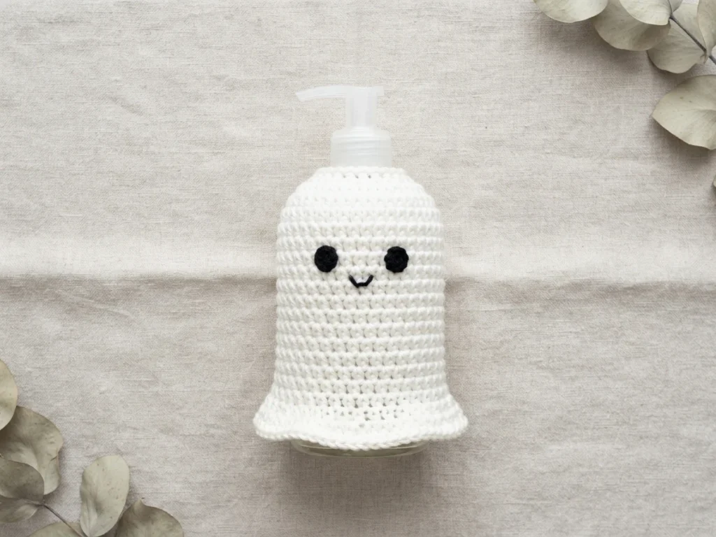White crocheted ghost soap cover with two black eyes and a small stitched smile, fitted over a clear soap dispenser pump on a light linen cloth, with dried eucalyptus leaves in the corners.