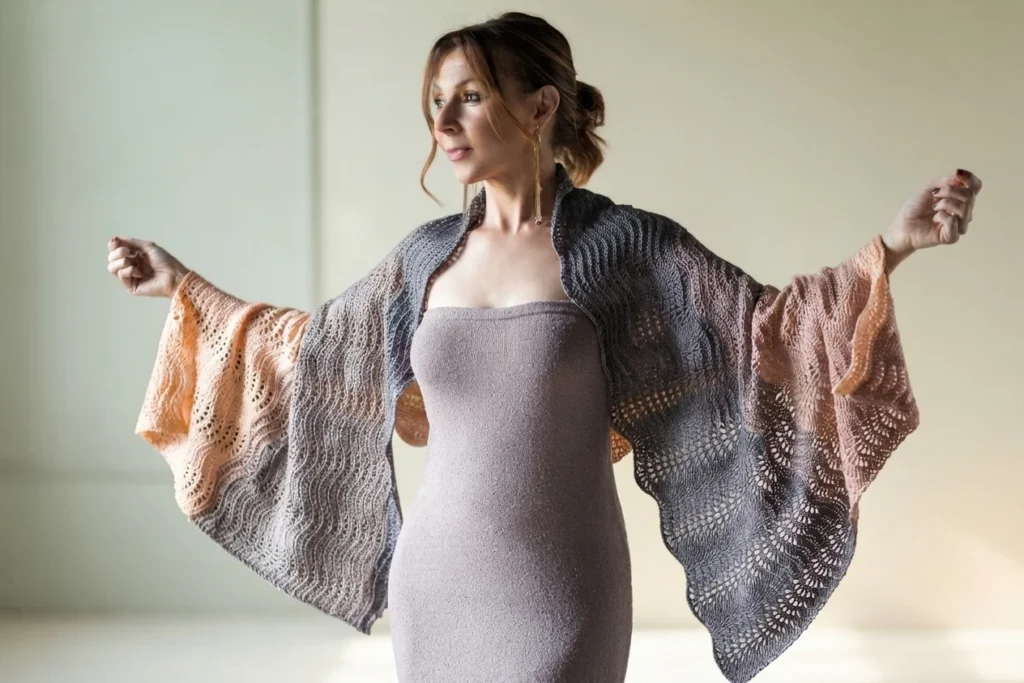 A woman wearing a gray and peach crocheted shawl with openwork patterns, holding her arms out to display it.