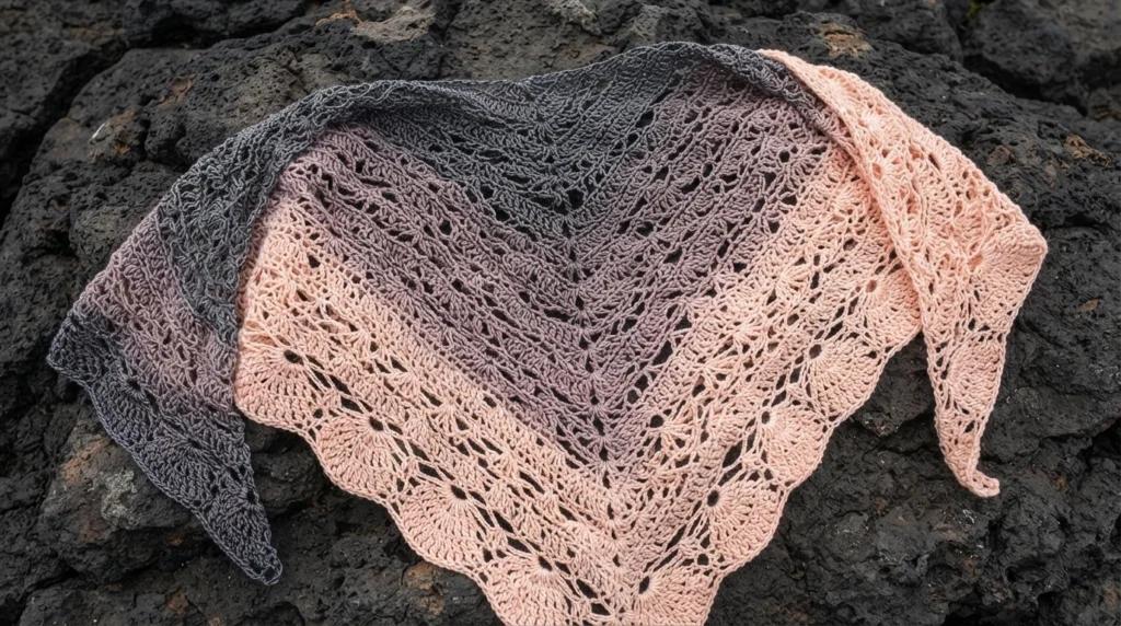 A lacy crochet shawl in charcoal gray, mauve, and peach, spread across dark rocks.