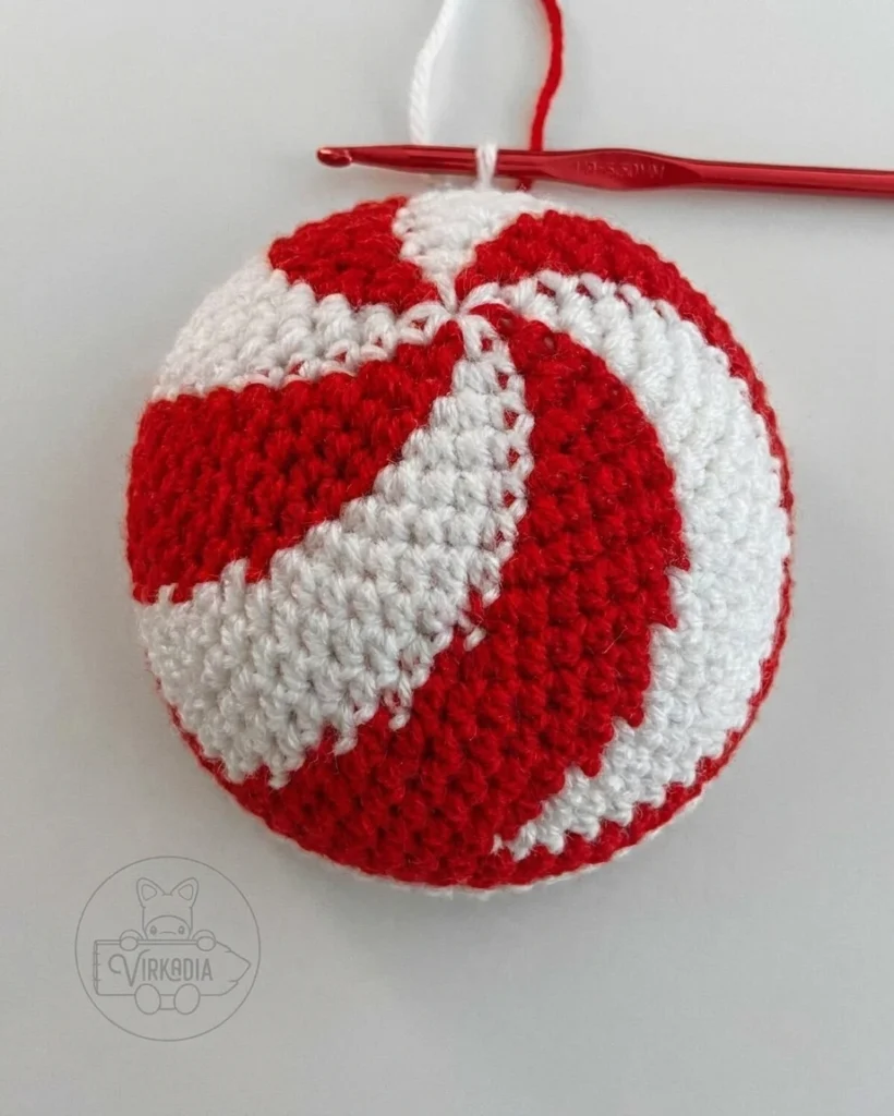 A red-and-white crocheted peppermint spiral crown growing in the round, with a red hook and working yarn.
